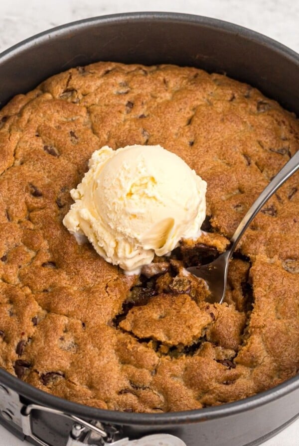 Golden cookie in a pan after being cooked in air fryer. Topped with vanilla ice cream and a spoon taking a scoop bit.