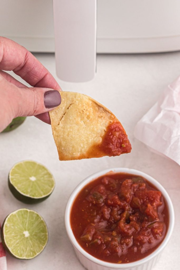 Hand holding a golden crispy tortilla chip dipped in salsa with slices of lime on the table
