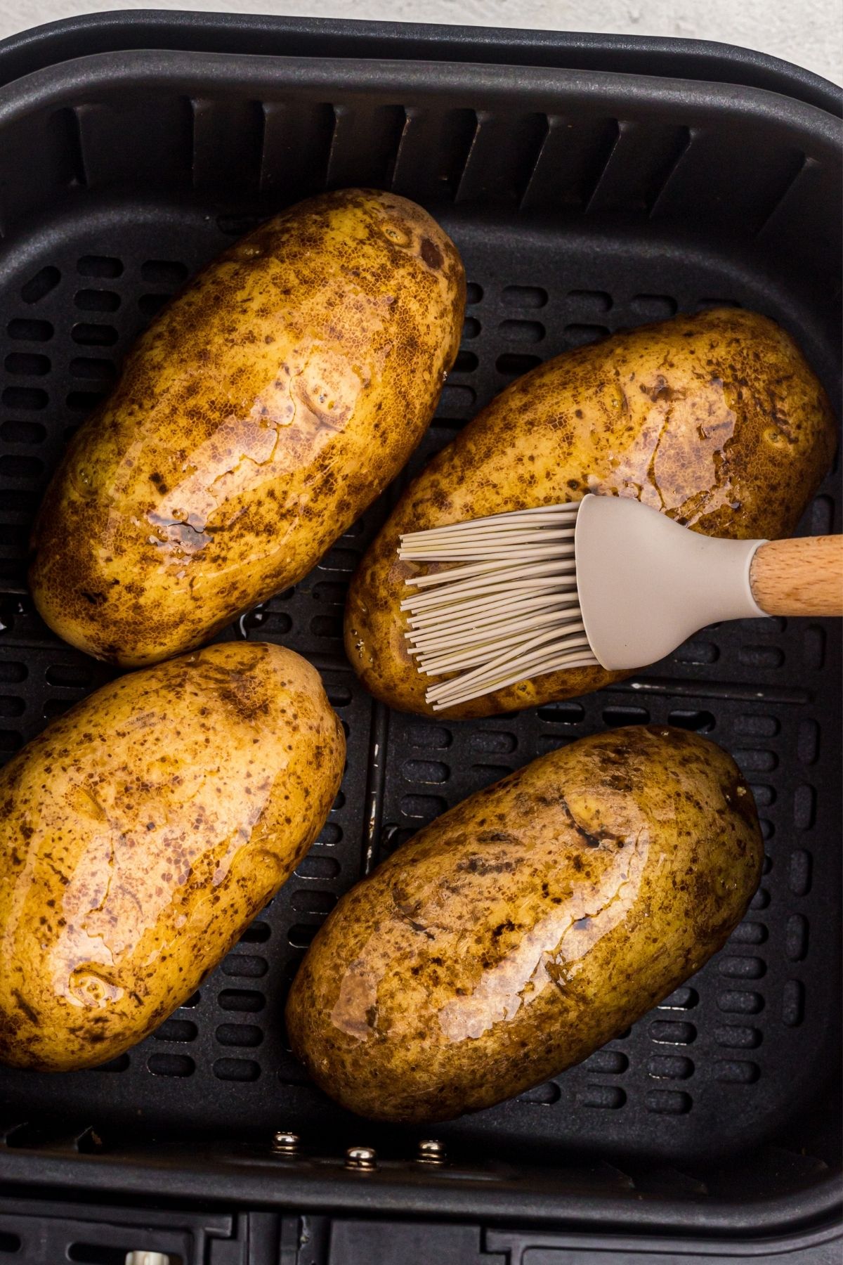 Four potatoes in the air fryer basket being brushed with olive oil