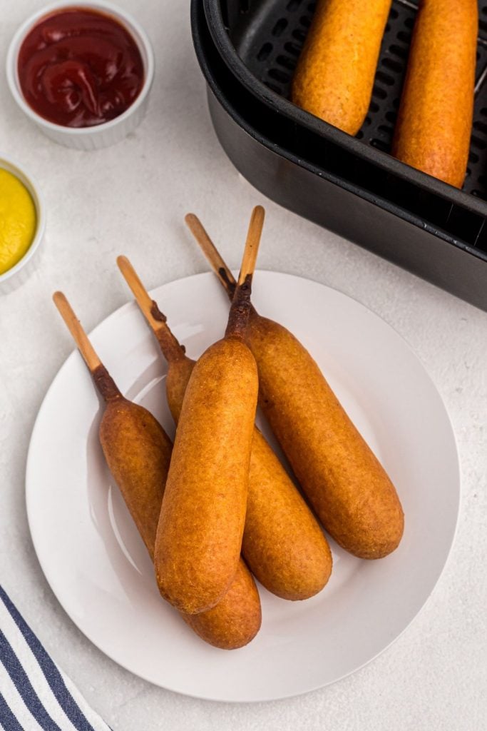 Golden corn dogs on a white plate with ketchup and mustard on the table. 