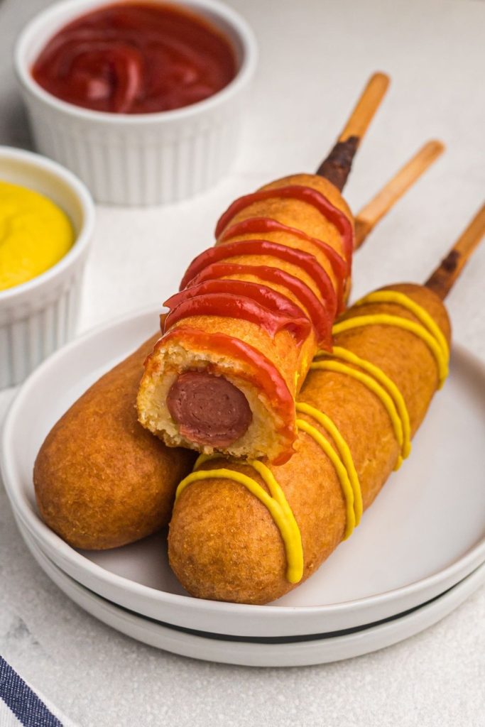Golden corn dogs on a small white plate with ketchup and mustard drizzled on top and a bite missing from one corn dog. 