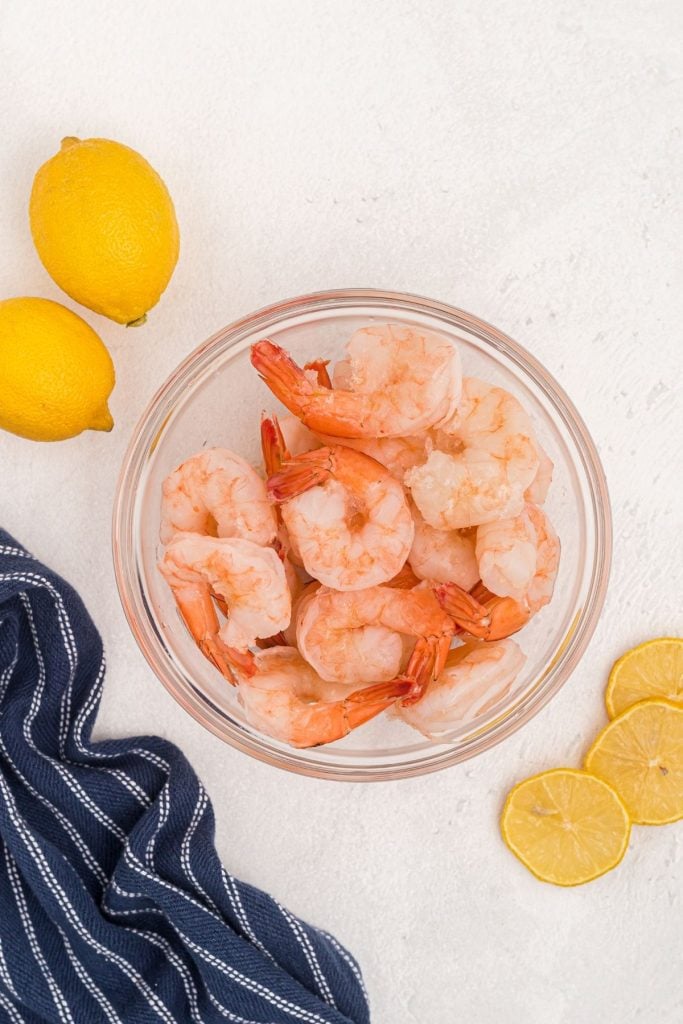 Frozen shrimp in a clear glass bowl with lemon slices on a white marble table. 