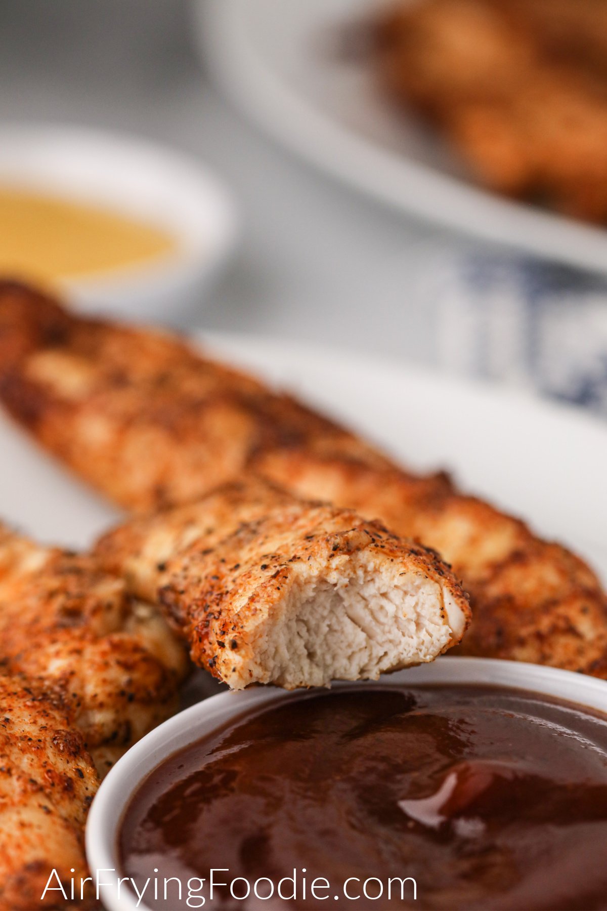 Chicken tender with a bite taken out of it over a small bowl of BBQ sauce. 