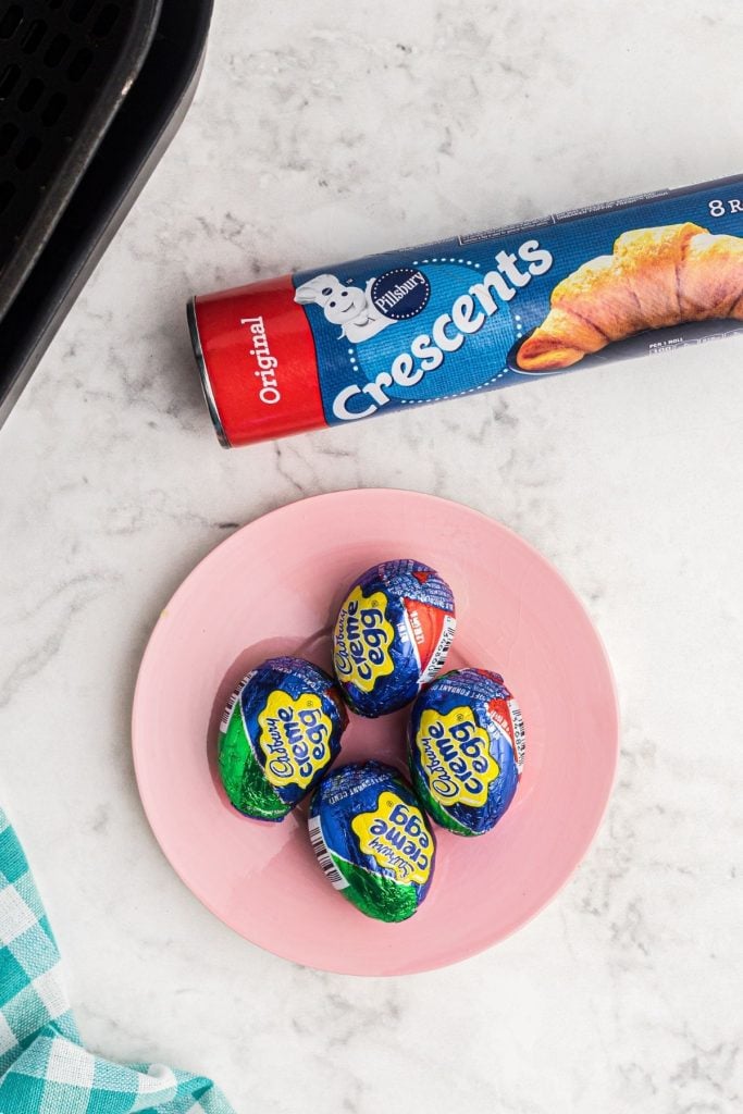 Ingredients needed to make fried cadbury eggs. Eggs on a pink plate and can of crescent rolls with air fryer basket. 

