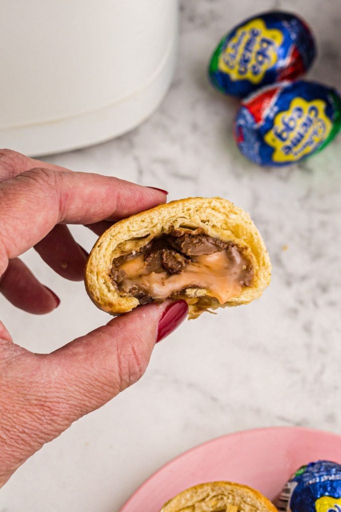 Gooey filling coming out of crescent wrapped chocolate egg being held by a hand in front of air fryer. 