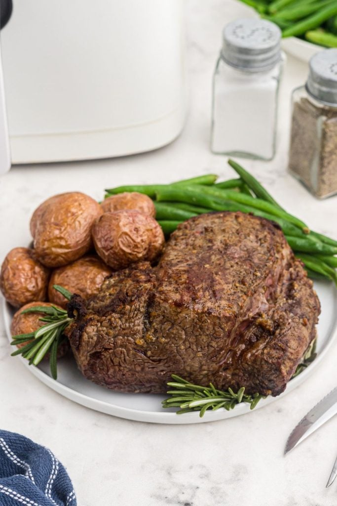Juicy brown beef tenderloin on a white plate with potatoes and green beans in front of the air fryer.
