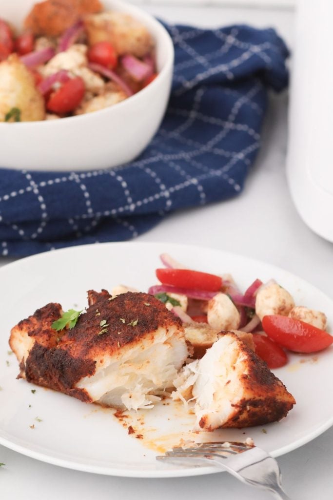 Flaky cod on a white plate seasoned and garnished with tomatoes and other veggies