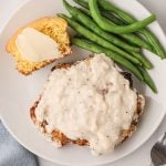 Overhead shot of chicken fried steak made in the air fryer and served on a plate covered in gravy and served with green beans and a side of cornbread.