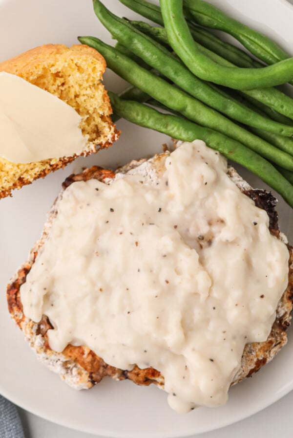 Overhead shot of chicken fried steak made in the air fryer and served on a plate covered in gravy and served with green beans and a side of cornbread.