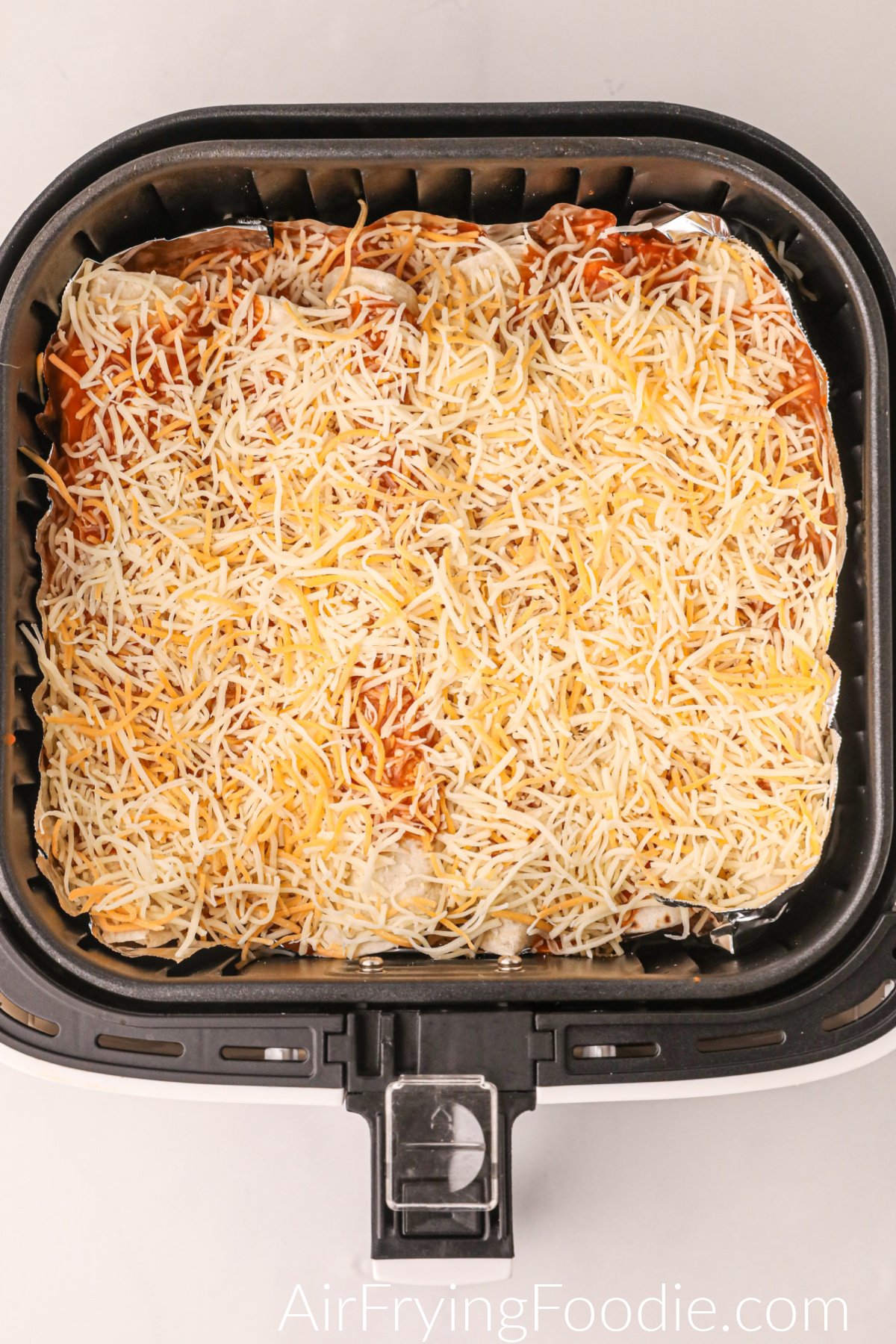 Enchiladas rolled into the bottom of the air fryer basket and topped with sauce and cheese, ready to air fry.