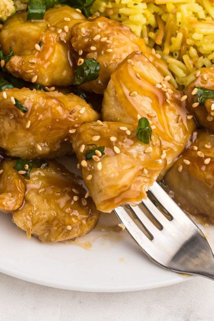 Close up bite of sweet and sour chicken on a fork, garnished with sesame seeds and green onions