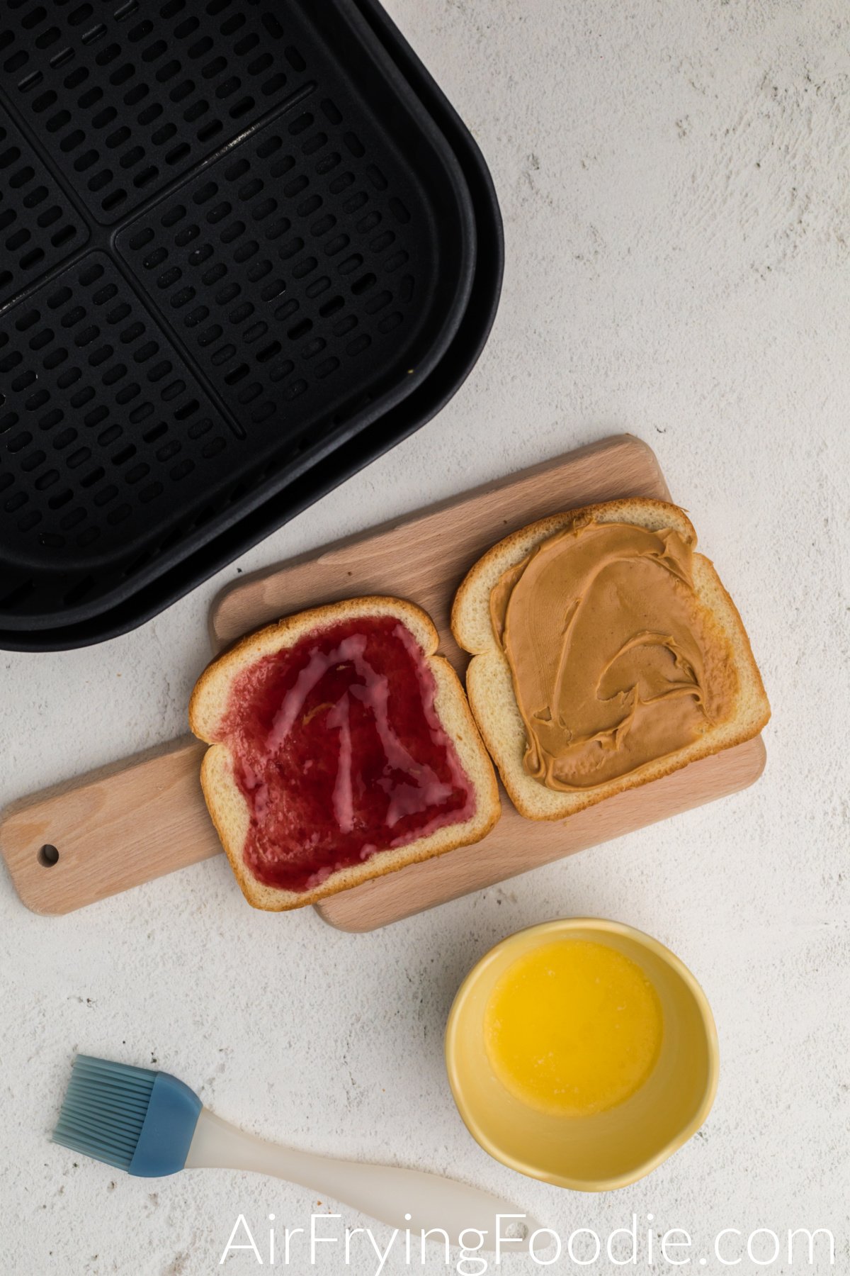Cutting board with bread that has peanut butter and bread with jelly, and a bowl of melted butter on the side.