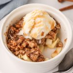 Apple crisp topped with oats and ice cream,drizzled with caramel sauce in a white ramekin