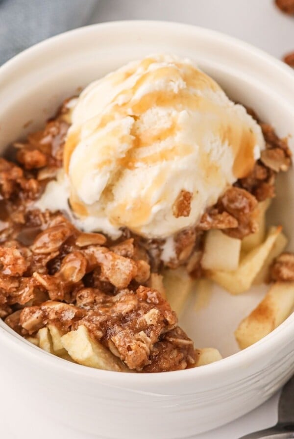 Apple crisp topped with oats and ice cream,drizzled with caramel sauce in a white ramekin