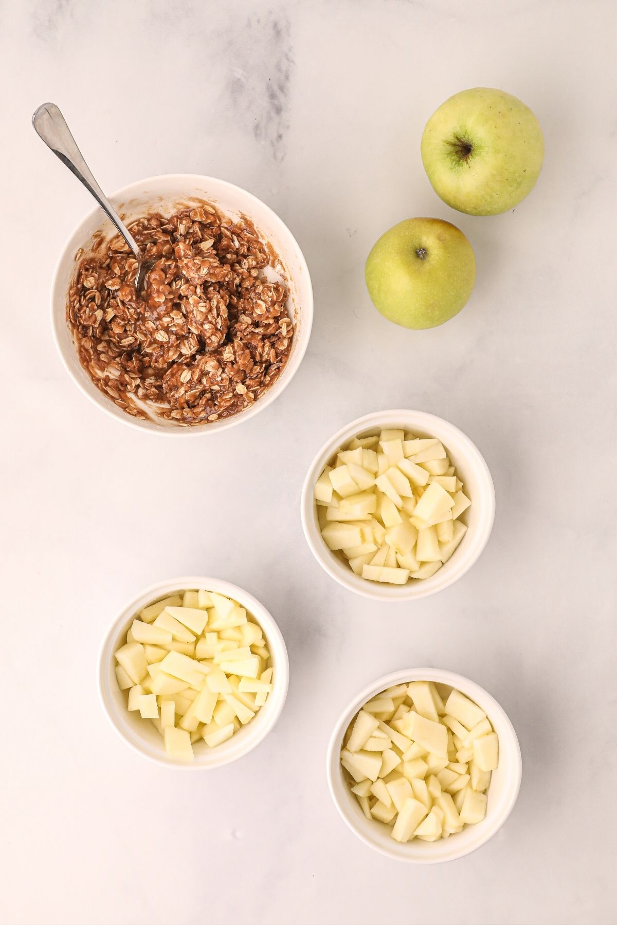 apples divided into ramekins and oats mixed with spices and sugars in a white bowl