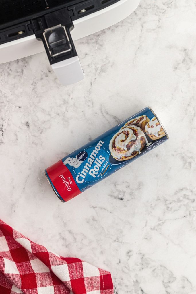 A can of cinnamon roll dough on a white marble table in front of an air fryer basket