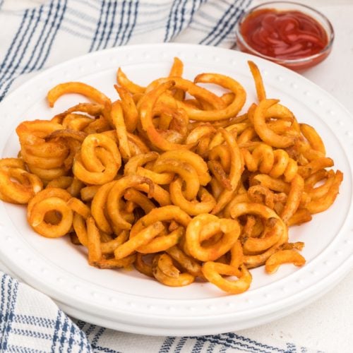 Air fryer frozen curly fries made and served on a white plate with a side of ketchup for dipping.