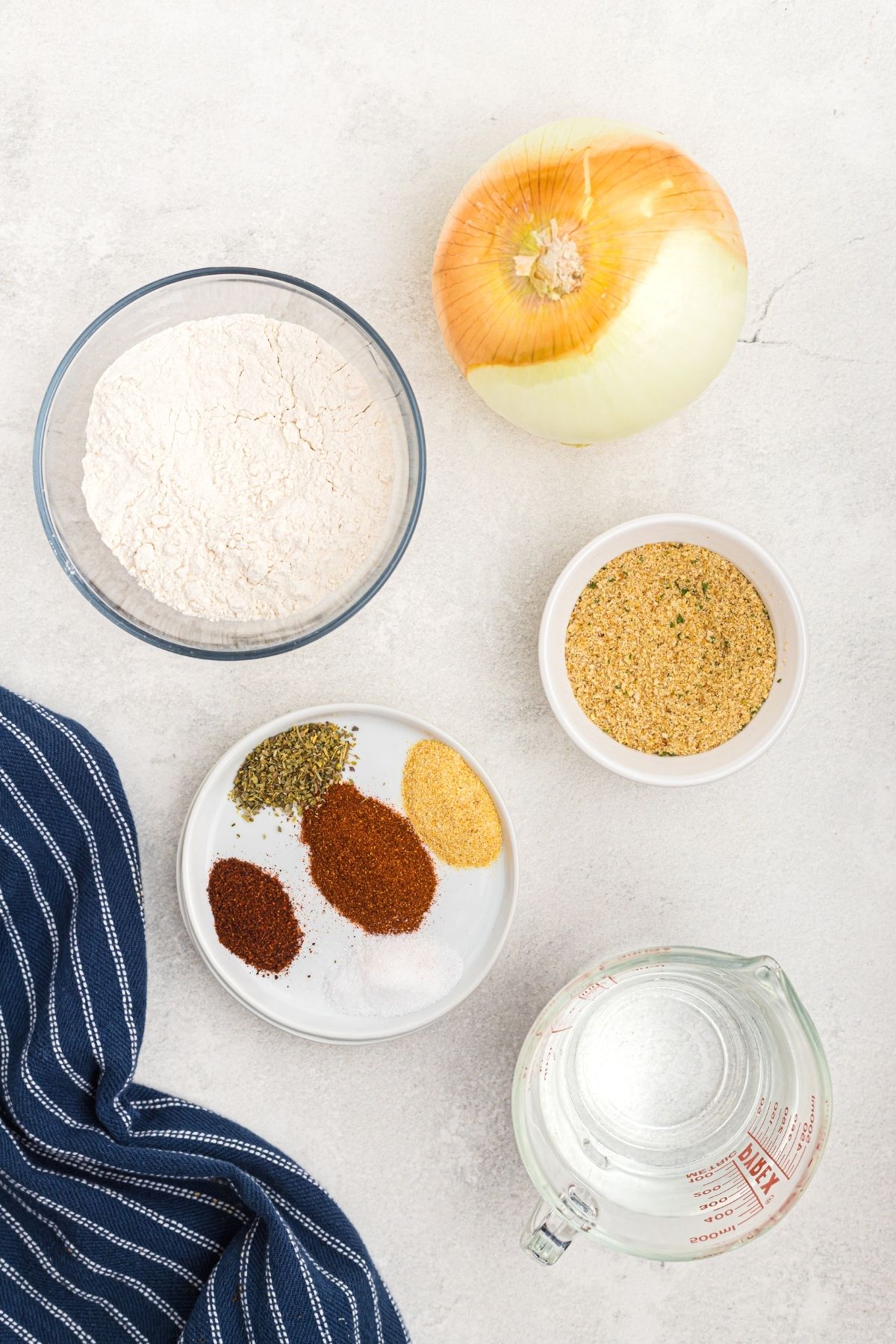 Crispy Air Fryer Blooming Onion 3 Ingredients for blooming onion: seasonings, breadcrumbs, whole onion, flour, laid out on a white marble board