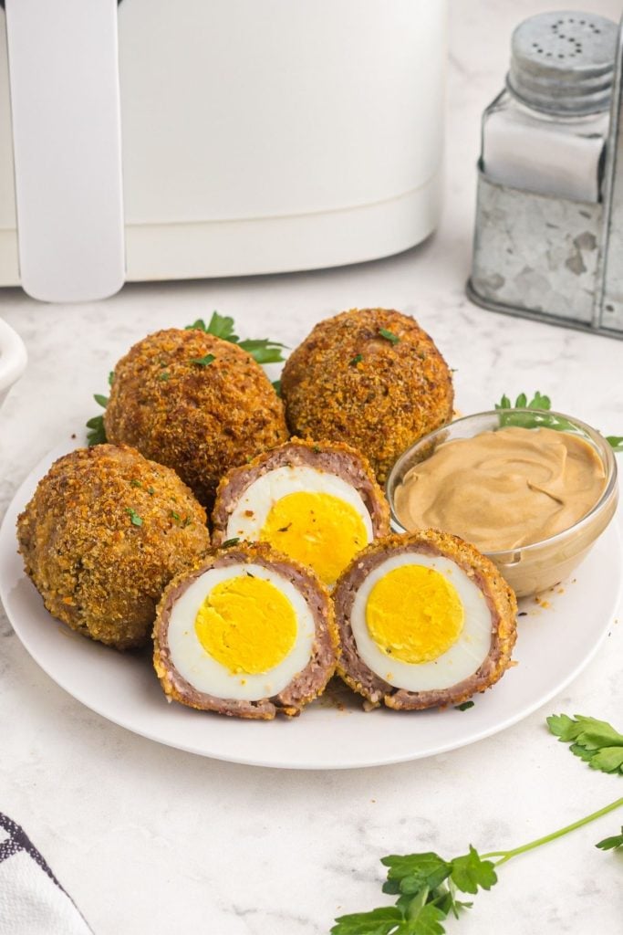 Coated hard boiled eggs with sausage and breadcrumbs, served on a white plate with Dijon mustard. 