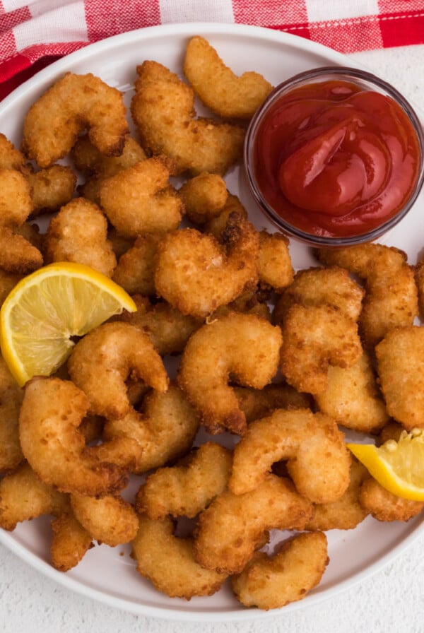 Overhead shot of golden brown frozen popcorn shrimp fully cooked in the air fryer and served on a white plate with dipping sauce and lemon wedges.