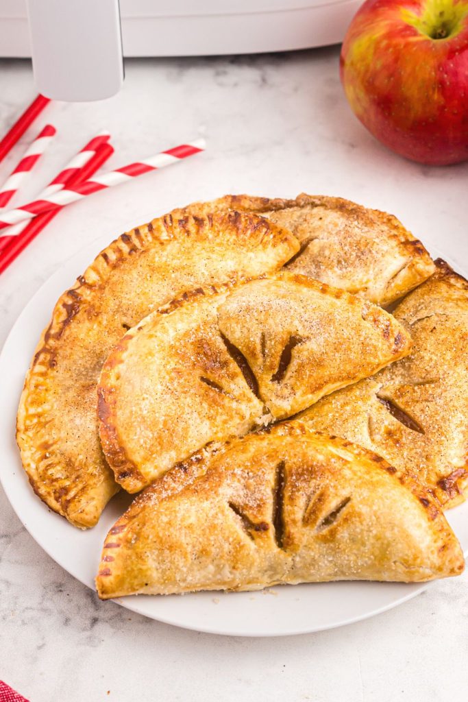 Golden crispy apple pies on a white plate in front of the air fryer with red straws and apples