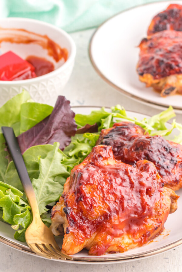 BBQ Chicken thighs on a plate with a side salad, and chicken thighs in the background with a bowl and basting brush with BBQ sauce.