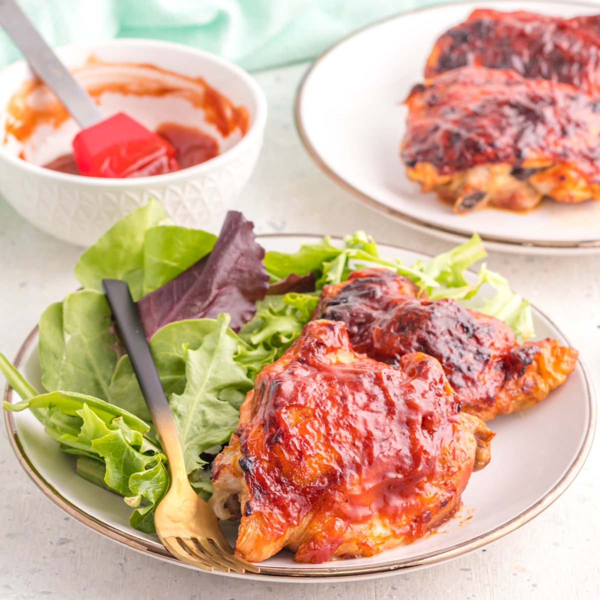 BBQ Chicken thighs on a plate with a side salad, and chicken thighs in the background with a bowl and basting brush with BBQ sauce.