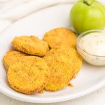 Fried Green Tomatoes on a white plate ready to serve with dipping sauce and a green tomato on the side.