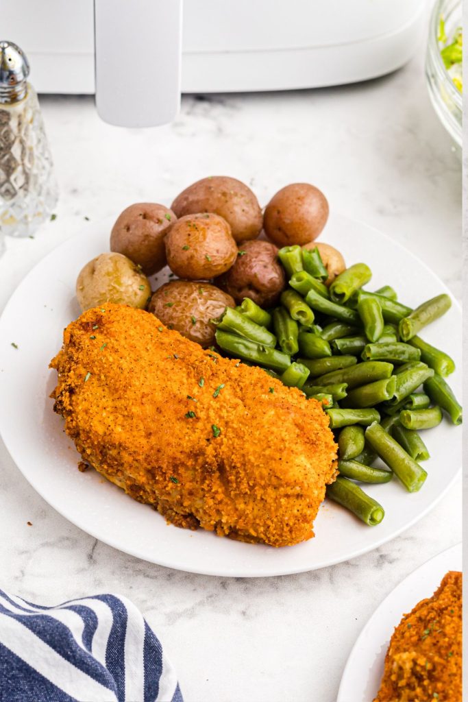 Golden crispy coated chicken breast on a white plate with green beans and potatoes.
