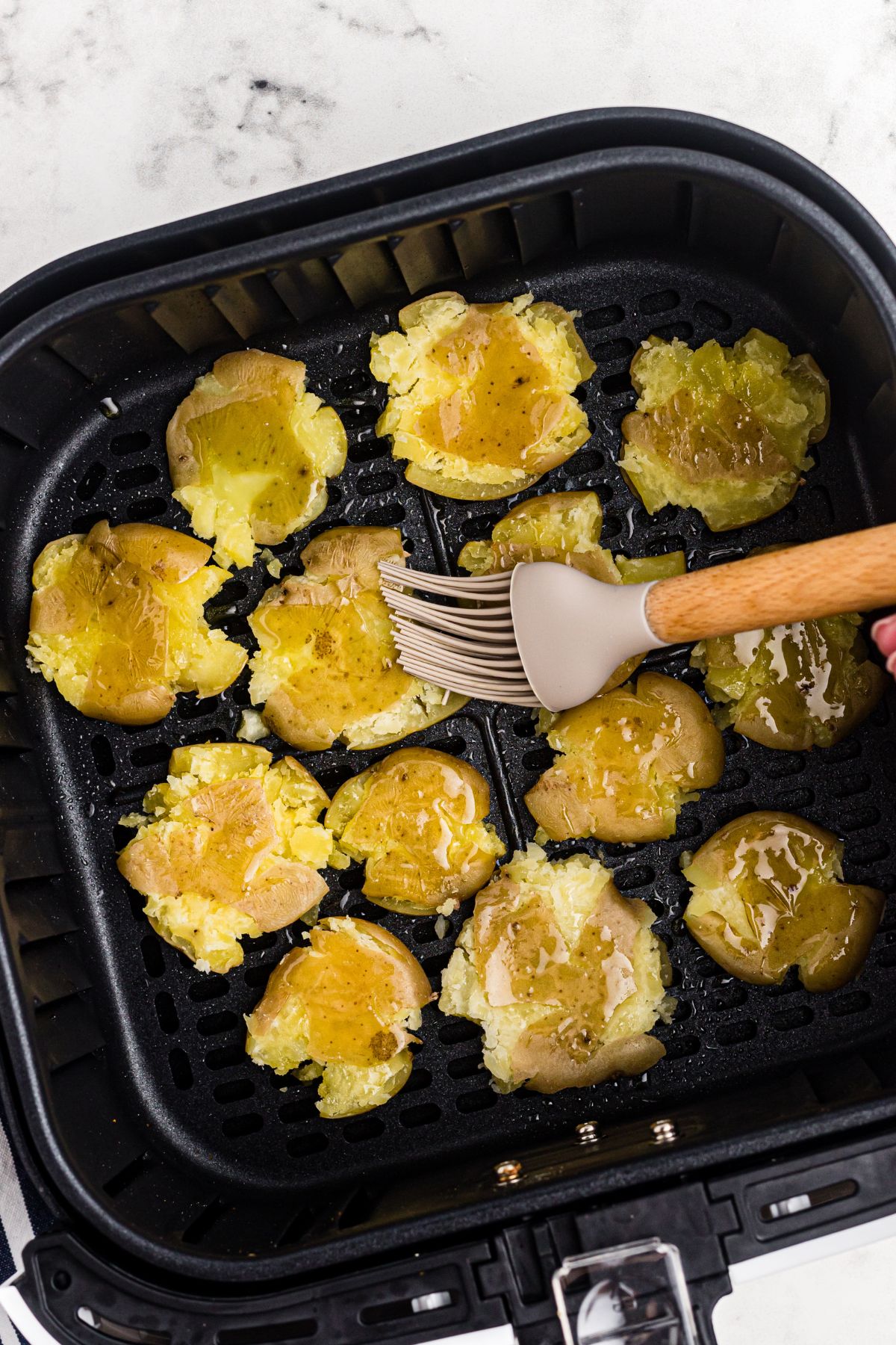 Smashed golden baby potatoes in the air fryer basket being brushed with olive oil and seasoned