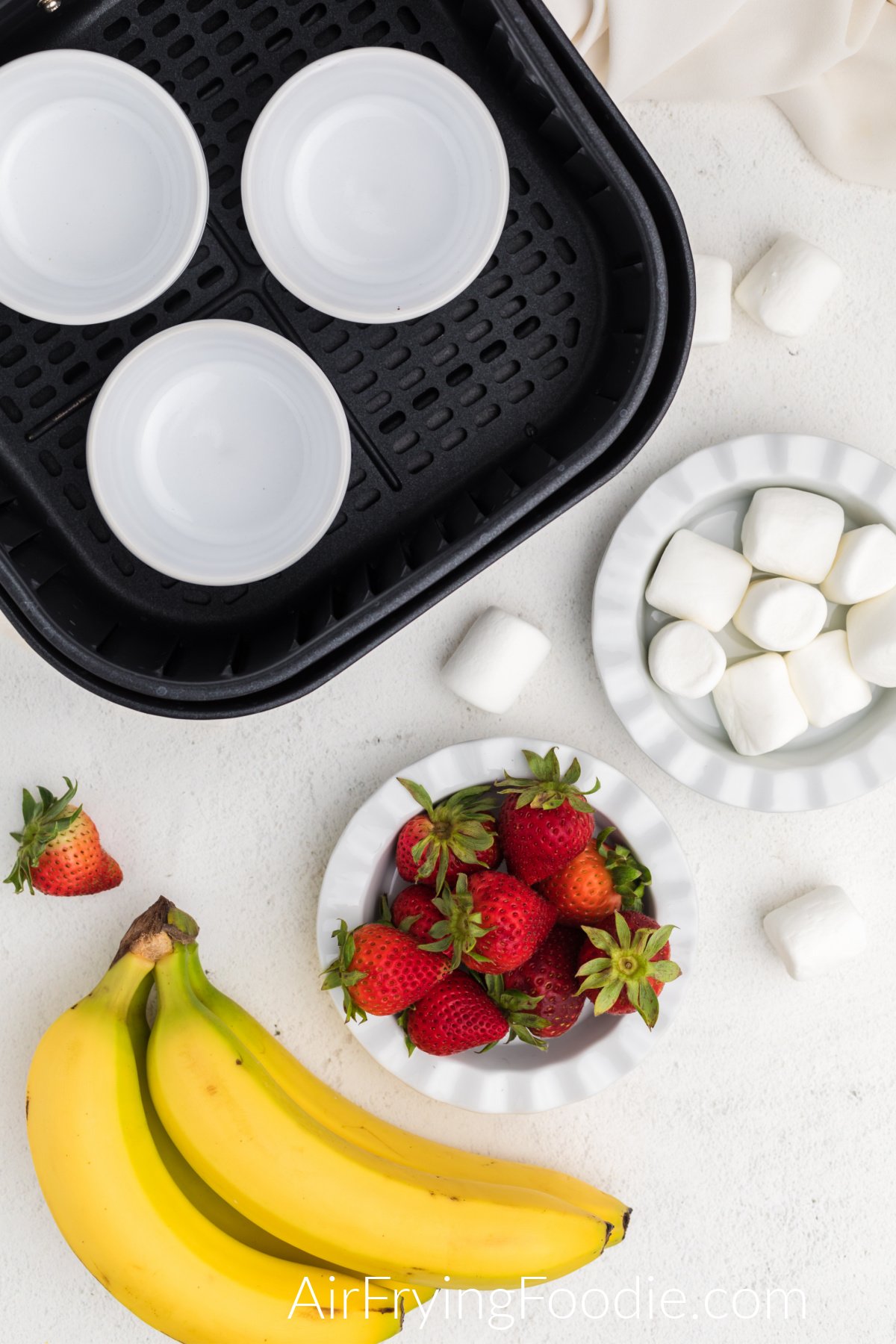 Air fryer basket with ramekins on a table with bananas, strawberries, and marshmallows.