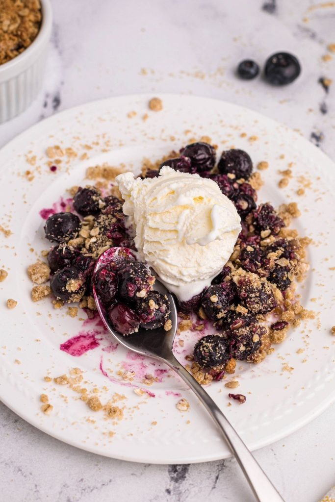 Blueberry crisp on white plate with blueberries on a spoon topped with vanilla ice cream.