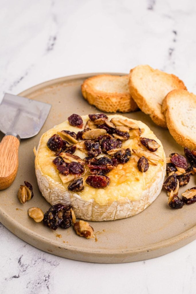 Wheel of brie cheese with nuts and berry toppings and toasted bread served on a tray.