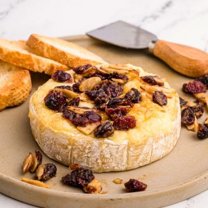 Wheel of brie cheese on a serving dish with nuts and berries and slices of bread on a platter.