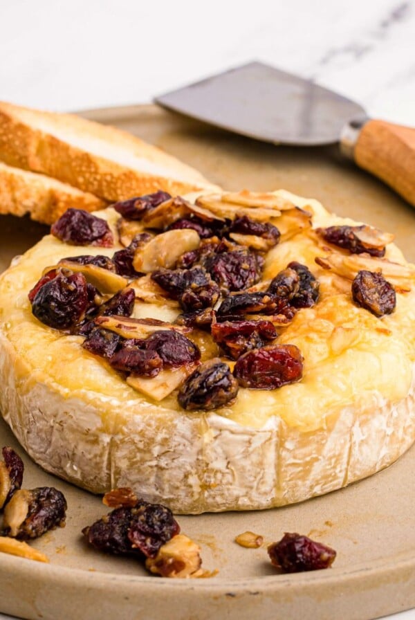 Wheel of brie cheese on a serving dish with nuts and berries and slices of bread on a platter.