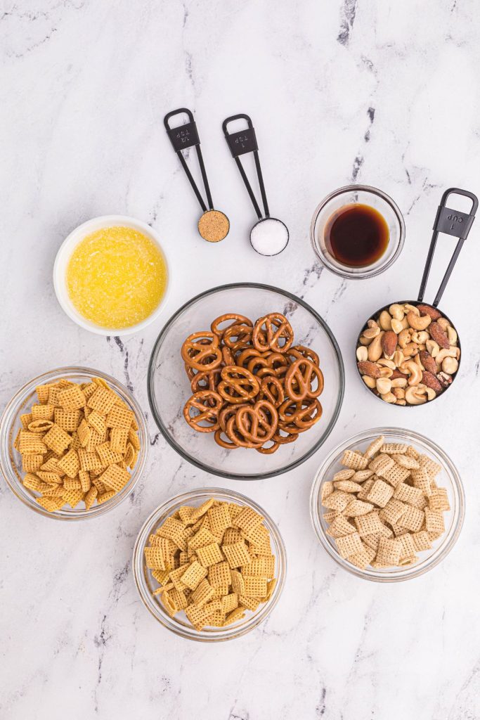 Ingredients needed for Chex mix on the table with pretzels, nuts, cereal, and others in measuring cups