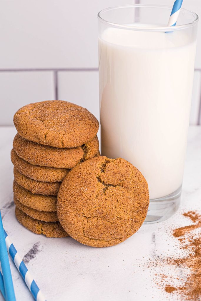 Golden gingersnaps coated in sugar and cinnamon stacked next to a large glass of milk with blue straws