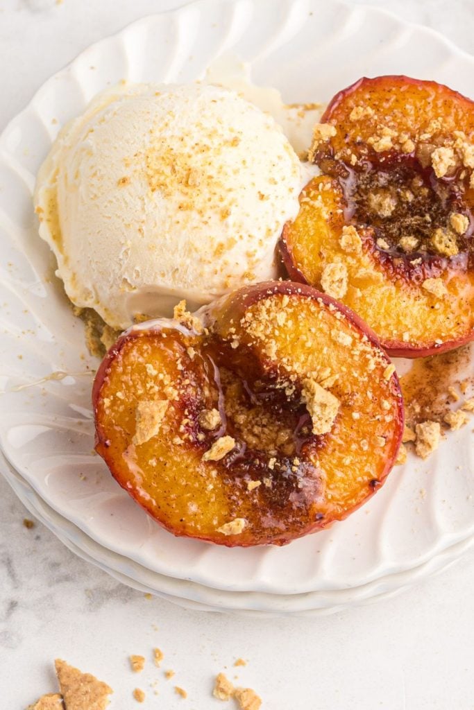 Juicy air fried peaches on a white plate served with a scoop of ice cream and topped with graham cracker crumbs