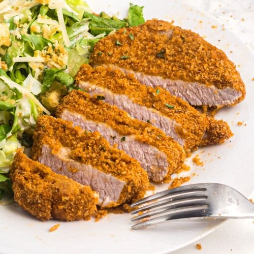 Golden crispy pork chop sliced on a white plate with salad and a fork