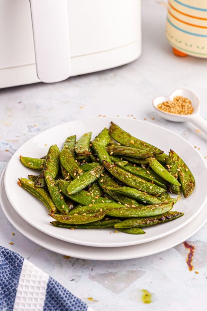 White plate with snap peas in front of an air fryer with sesame seeds scattered on the table. 