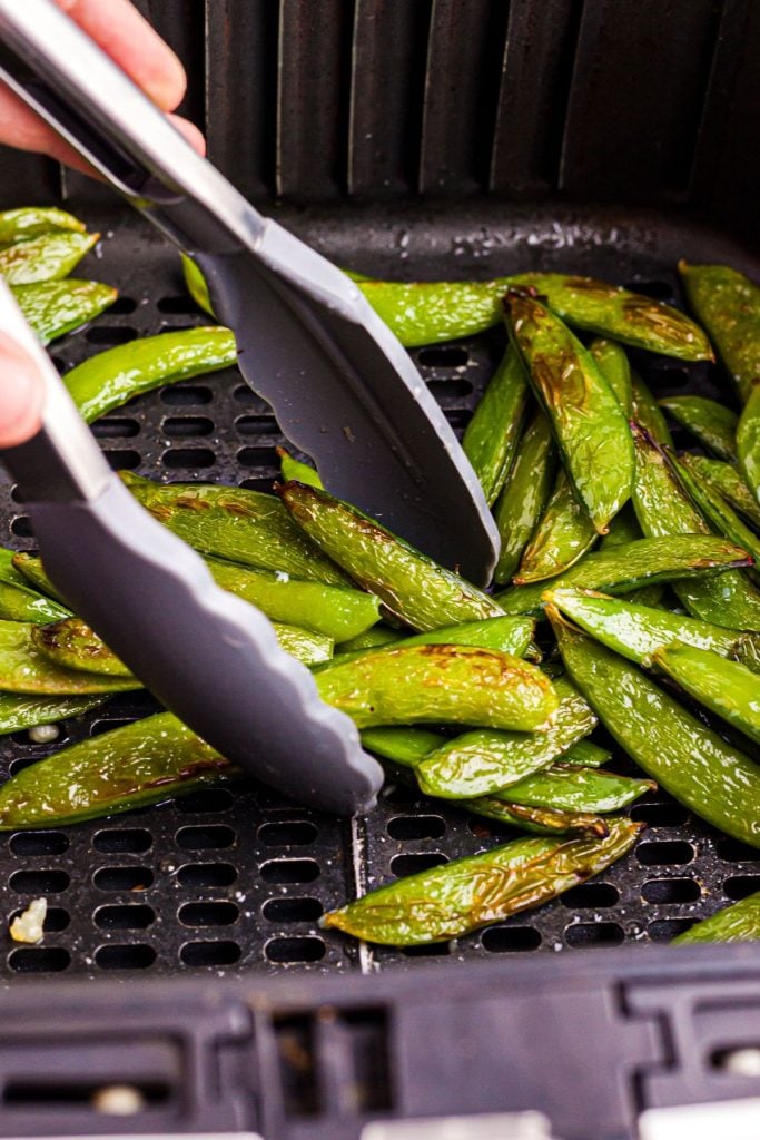 Air fried snap peas in the air fryer basket after being cooked , being served with tongs