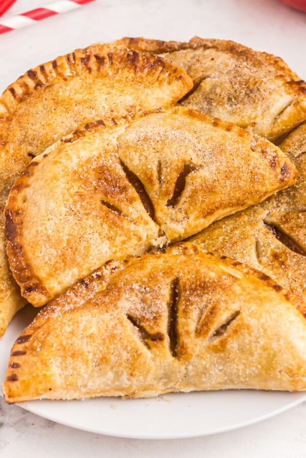 Golden hand held apple pies stacked on a white plate with red straws scattered on table.