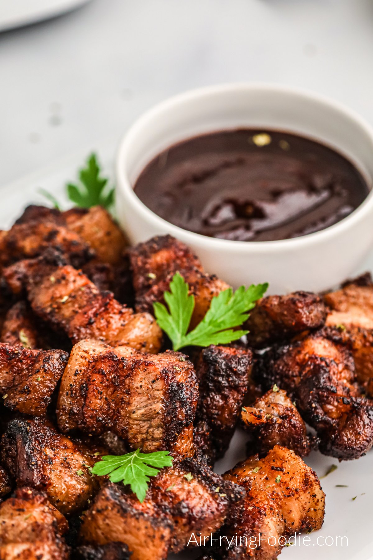 Pork belly bites on a white plate with fresh parsley and BBQ dipping sauce. Ready to serve.