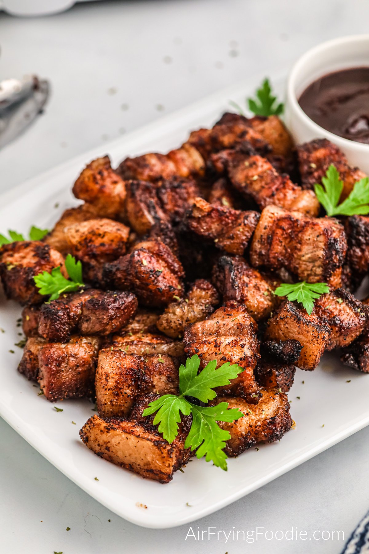 Air fryer pork belly bites in a plate with fresh parsley and BBQ dipping sauce.
