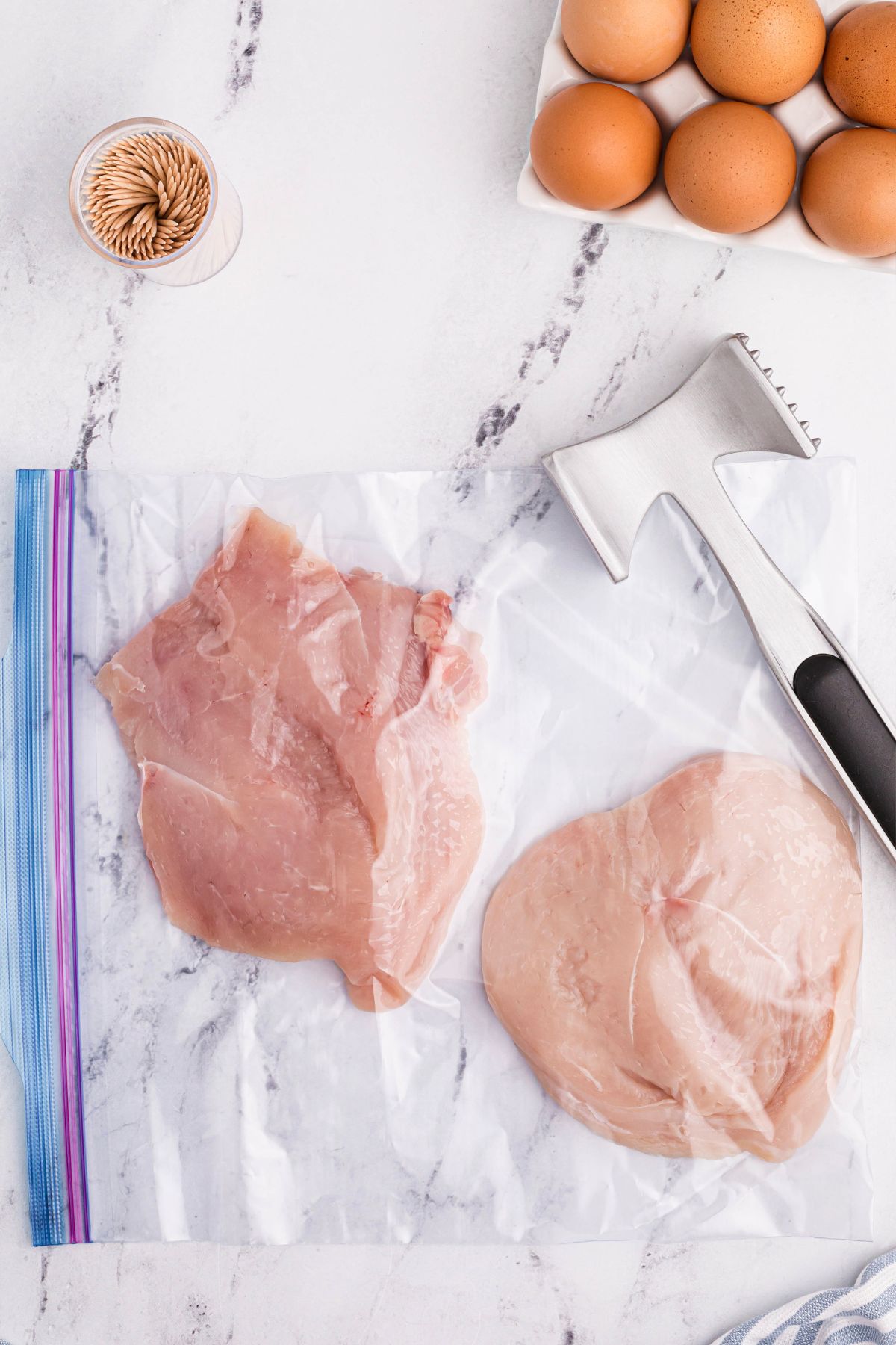raw chicken in a large Ziploc bag with a meat tenderizer on the table to flatten them