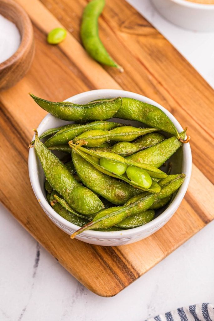 Bright green edamame pods in a white bowl seasoned on a brown board.