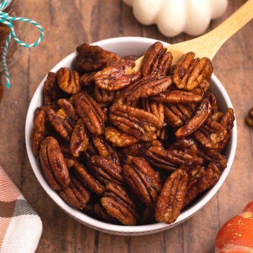 Juicy brown pecans in a white bowl after being cooked in the air fryer with scattered pecans on a wooden table