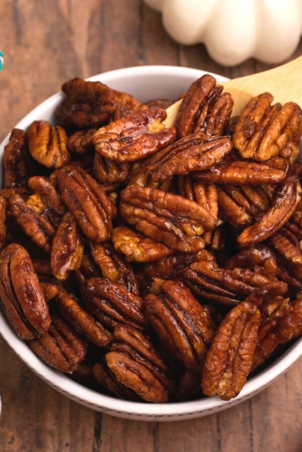 Juicy brown pecans in a white bowl after being cooked in the air fryer with scattered pecans on a wooden table