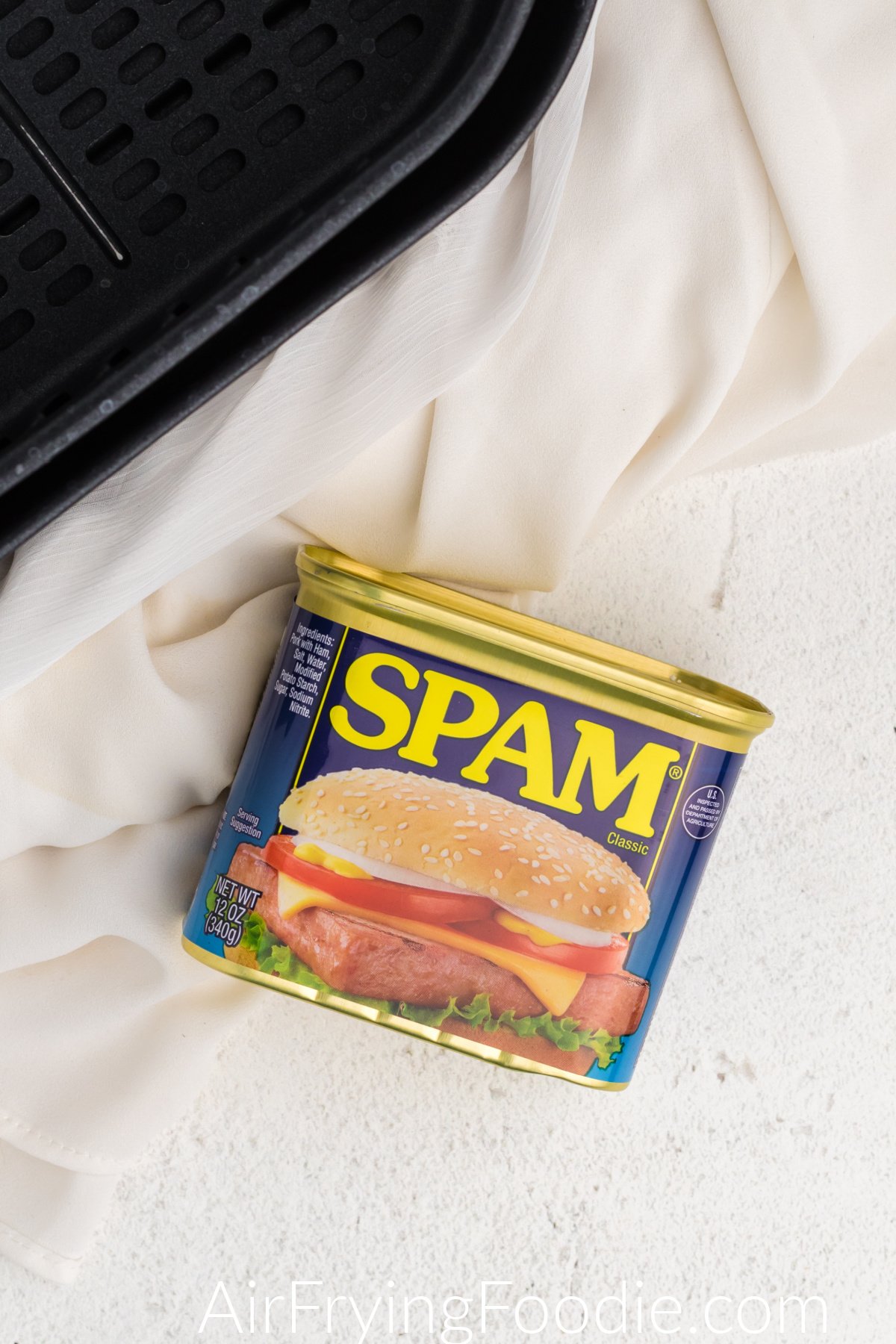 Can of classic Spam and the basket of the air fryer on a white table.