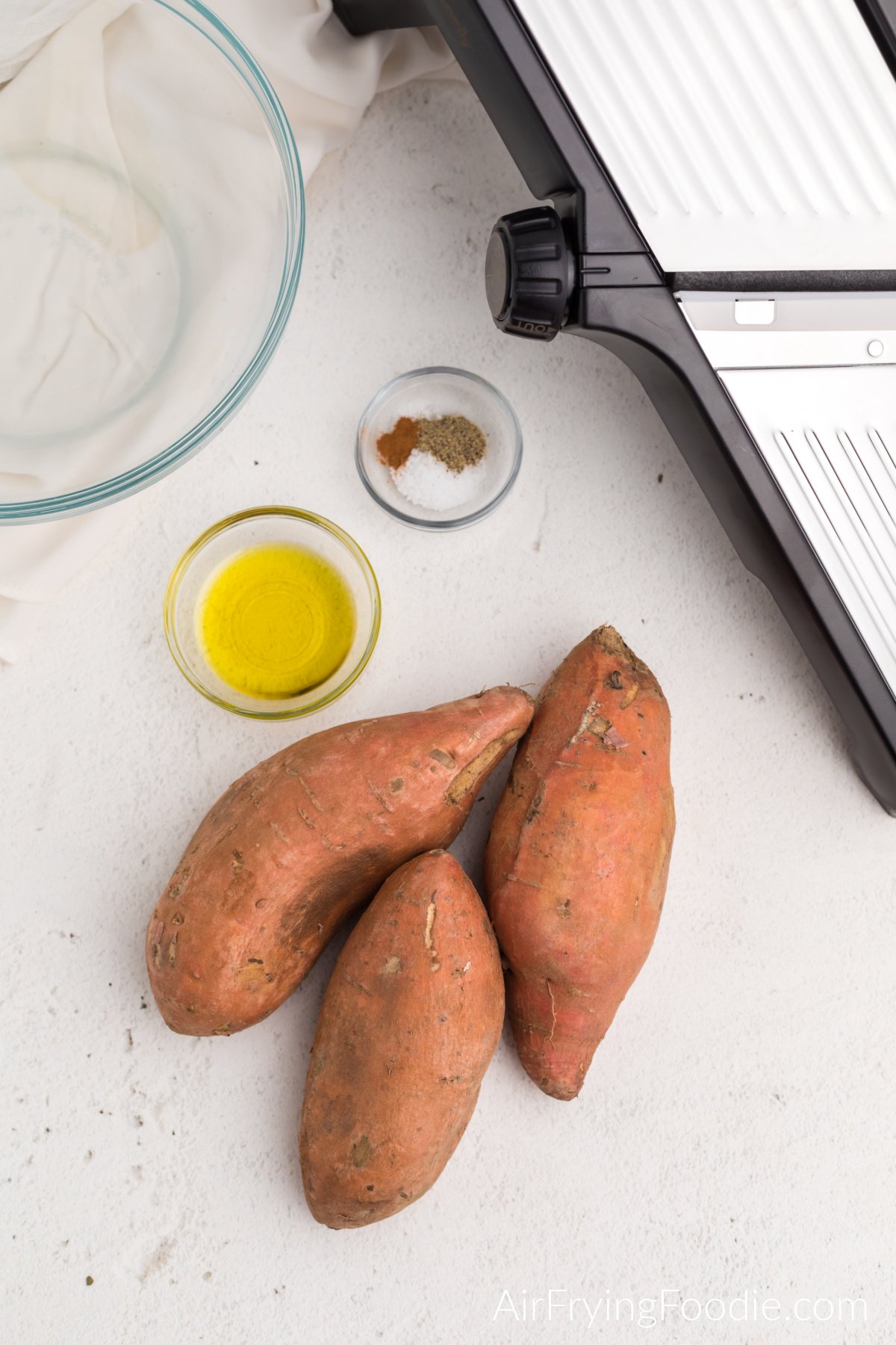 Olive oil, seasonings, sweet potatoes, a glass bowl, and a mandolin on a table. 